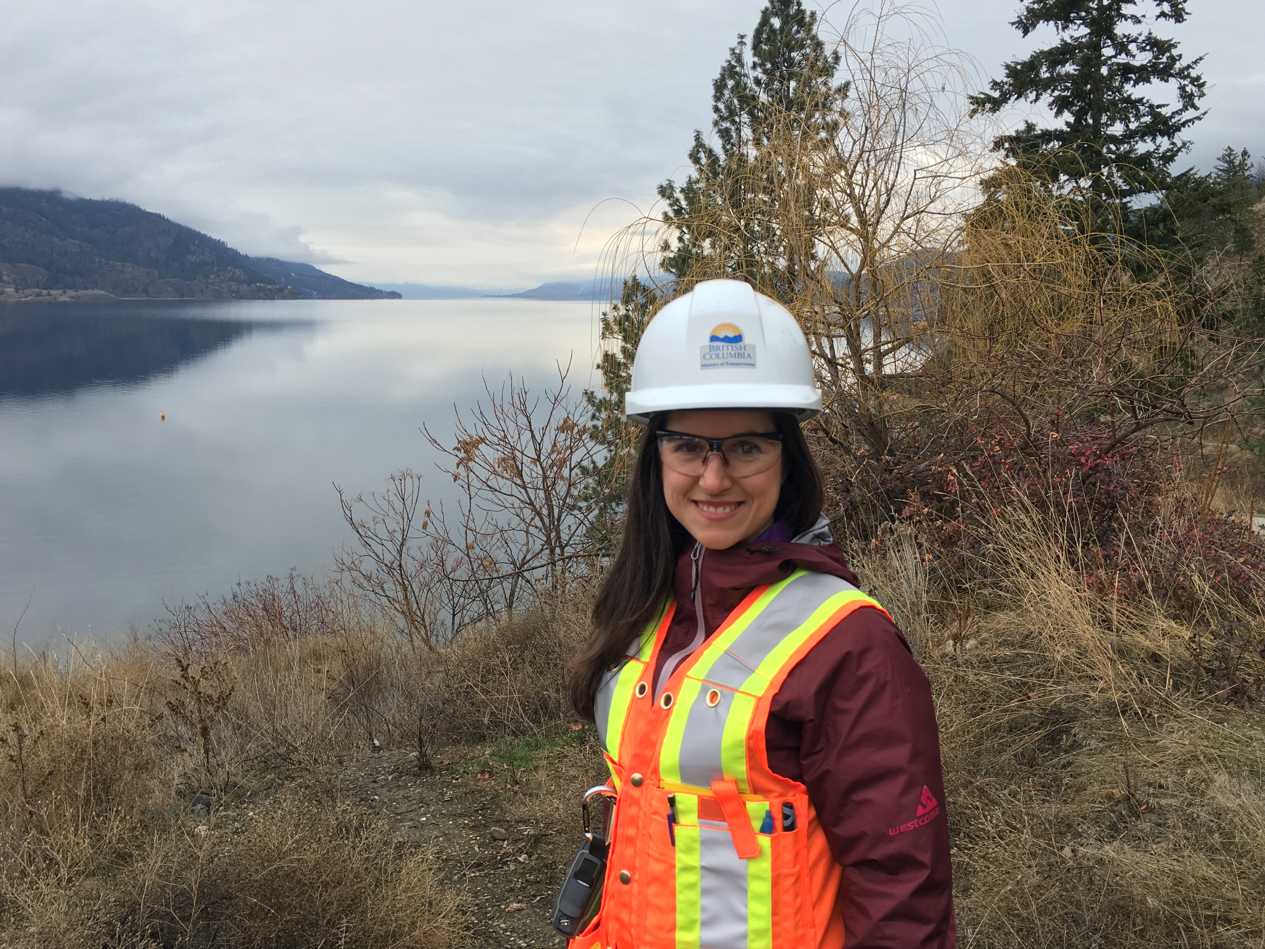 Female Geoscientist in orange reflective vest and hardhat looking into the camera with a lake and forest vegetation behind her