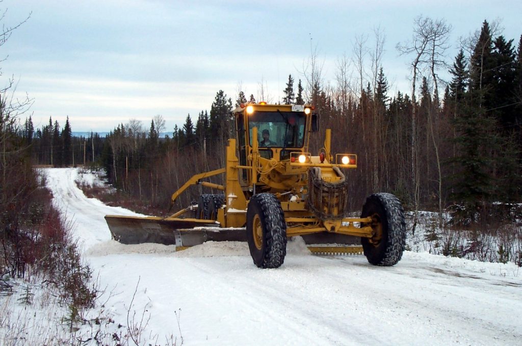 A grader with an under body plow working on clearing a rural road.